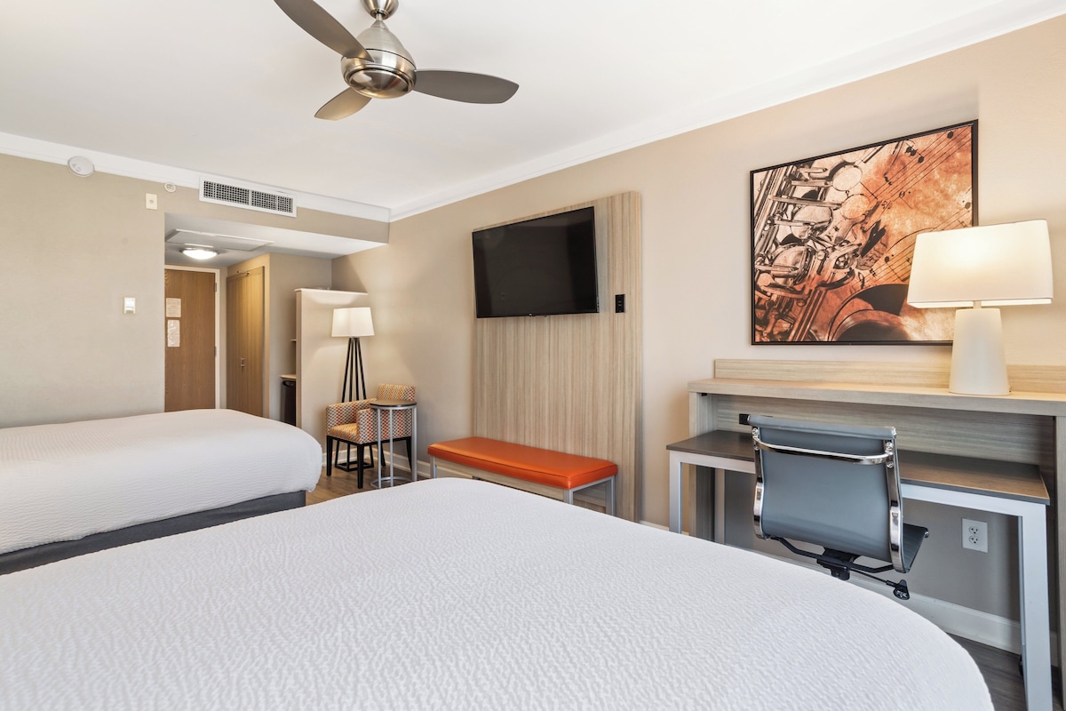 Ein Hotelzimmer mit zwei Betten, einem Schreibtisch mit Stuhl, einem Fernseher und einem Bild an der Wand.