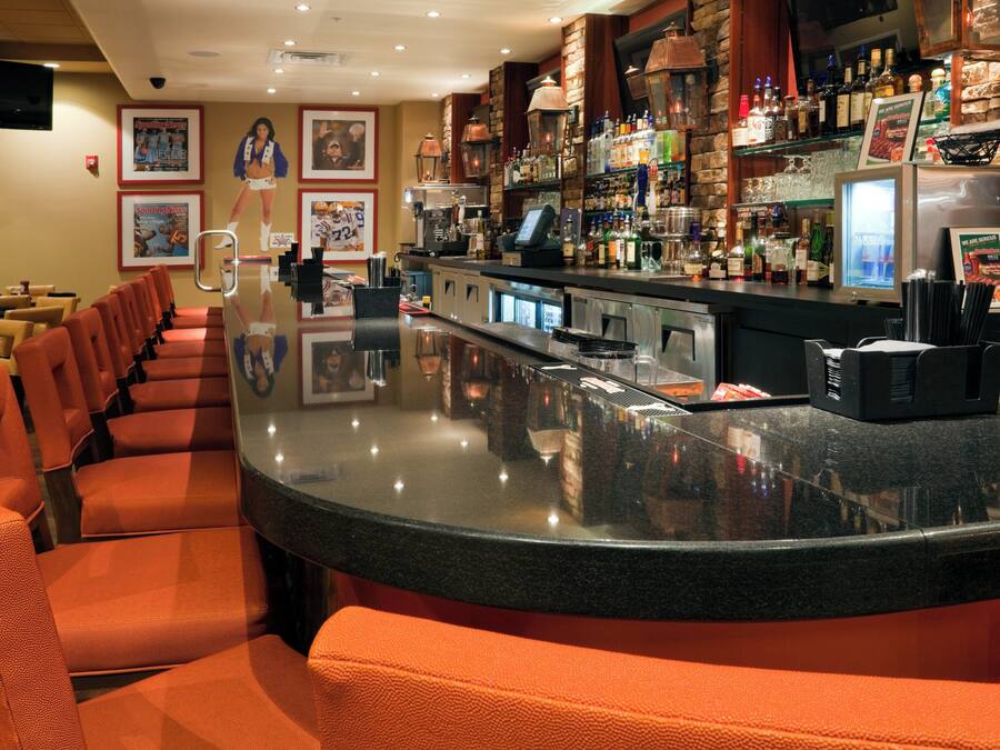 A bar area with a curved counter, seating with orange cushions, and a variety of bottles and glasses on shelves.
