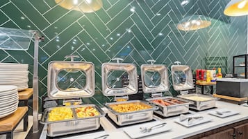 A buffet with various food items in metal containers, a stack of white plates, and condiment bottles on the counter.