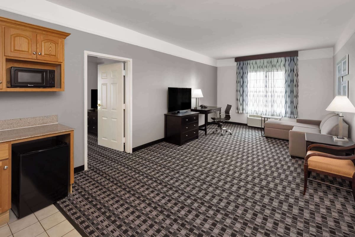 A hotel room with a kitchenette, a flat-screen TV, a sofa, a desk, and a patterned carpet.