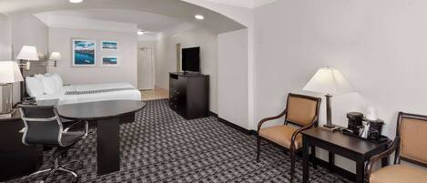 Quarto de hotel com cama, escrivaninha, cadeiras, televisão e pequena mesa com abajur.