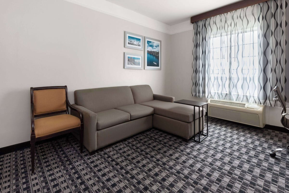 A modern hotel room with a sectional sofa, a single armchair, a small table, and a large window with patterned curtains.