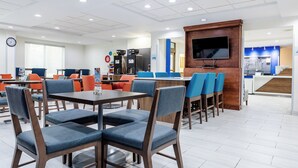 A modern hotel lobby with multiple dining tables and chairs, a coffee machine, and a TV mounted on a wooden panel.