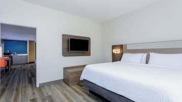 A hotel room with a bed, a TV mounted on the wall, a desk with a chair, and a kitchenette in the background.