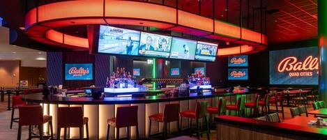 Zona de bar con varias pantallas mostrando eventos deportivos, una barra con taburetes y un gran letrero iluminado que dice "Bally's Quad Cities".