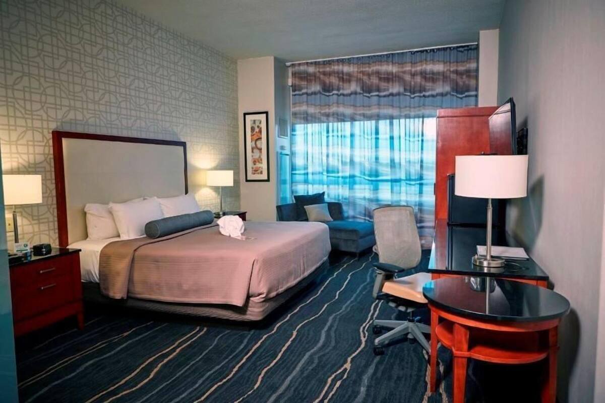 A hotel room with a large bed, a desk, a chair, a TV, and a window with curtains.
