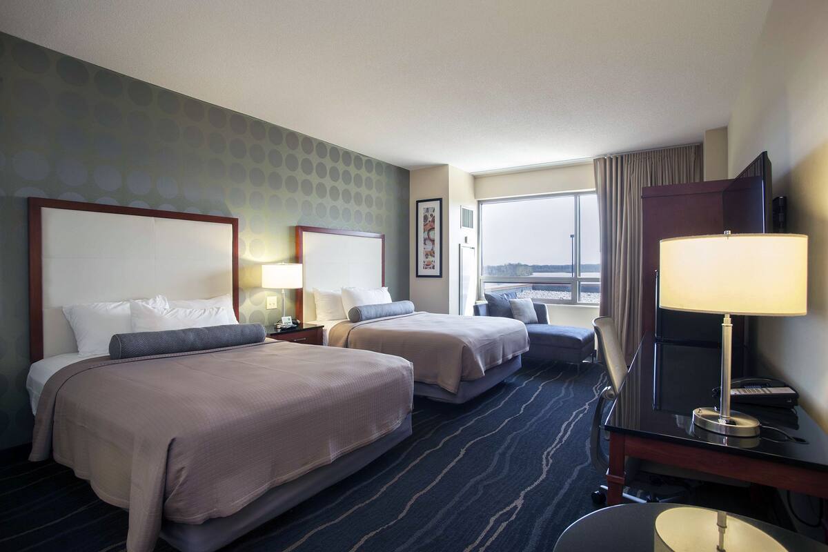 A hotel room with two beds, a desk with a lamp, a chair, and a large window offering a view.