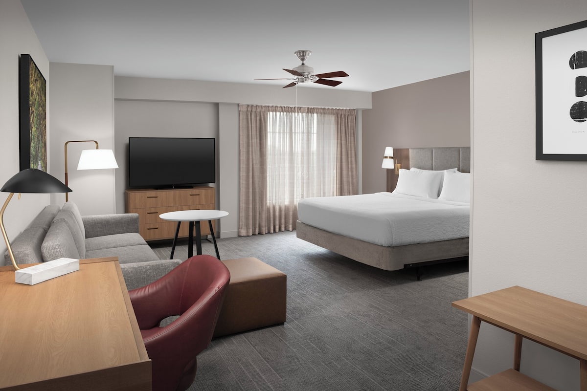Una habitación de hotel con cama, escritorio con lámpara, televisor, sofá y ventilador de techo.
