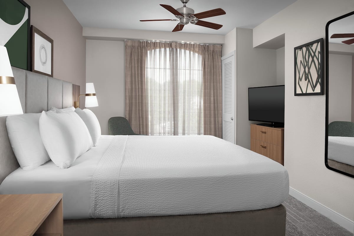 Habitación de hotel con una cama grande, ventilador de techo, televisor y un espejo en la pared.