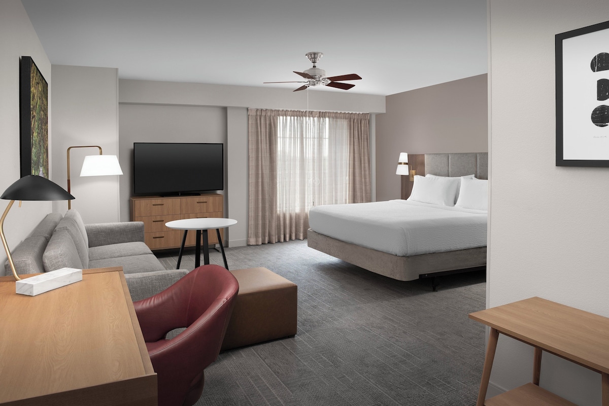 A hotel room with a bed, a desk with a lamp, a TV, a sofa, and a ceiling fan.