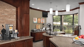 A kitchen with a brick wall, wooden cabinets, a coffee machine, a waffle maker, and a large window with a view of trees. - Staybridge Suites Columbia by IHG (Columbia)
