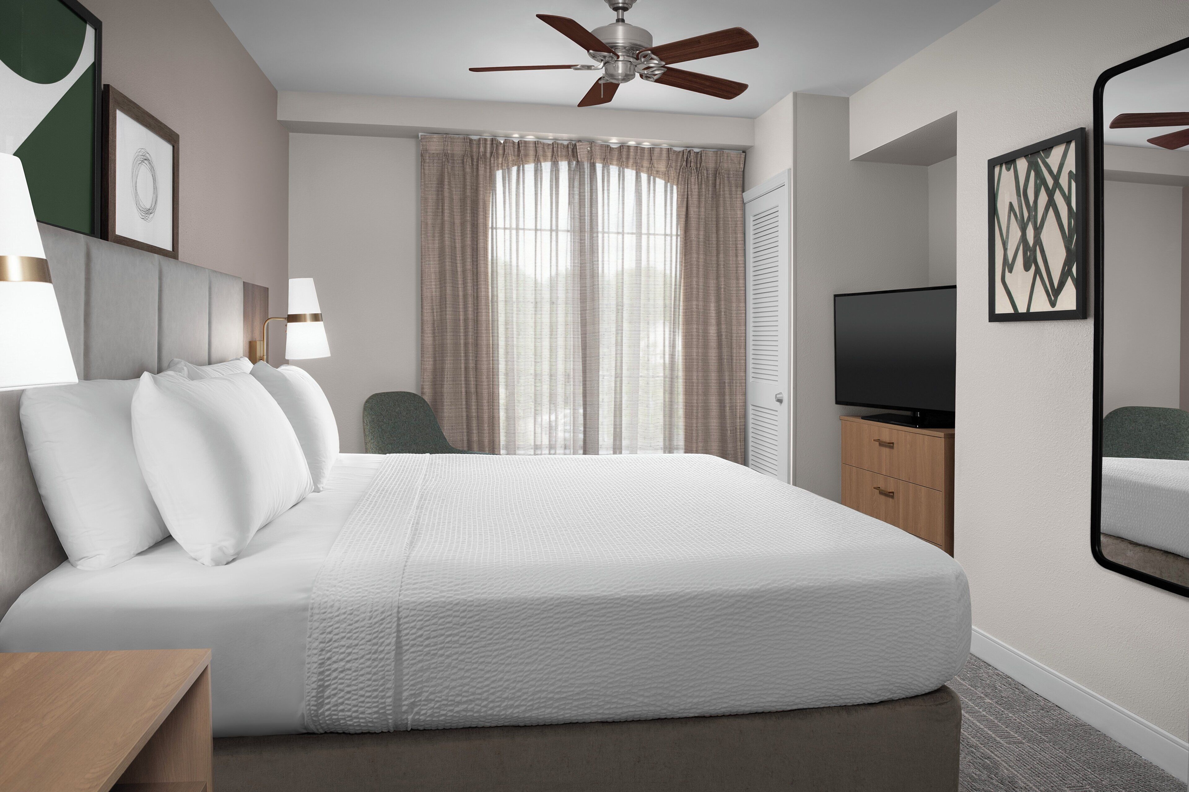 A hotel room with a bed, a ceiling fan, a TV, a mirror, and a window with curtains.