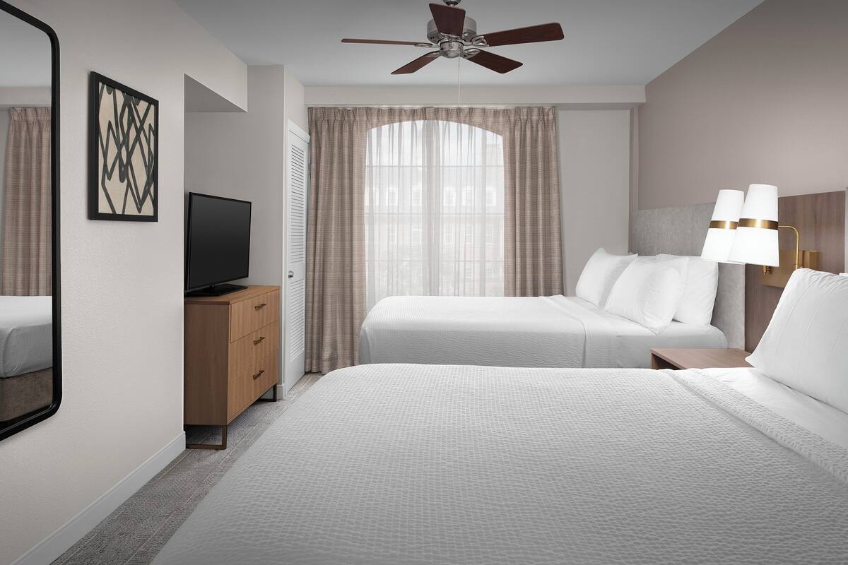 Una habitación de hotel con una cama, televisión, un mueble de madera, ventilador de techo y ventana con cortinas.