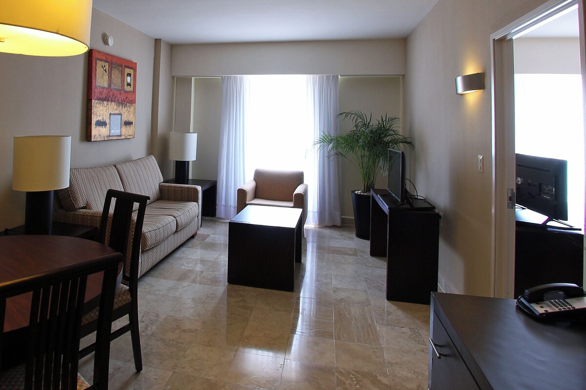 A hotel room with a sofa, a coffee table, a dining table with chairs, a TV, a desk with a phone, and a potted plant.