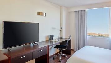 A hotel room with a bed, a desk with a television, a chair, a coffee maker, and a window with a view of the desert.