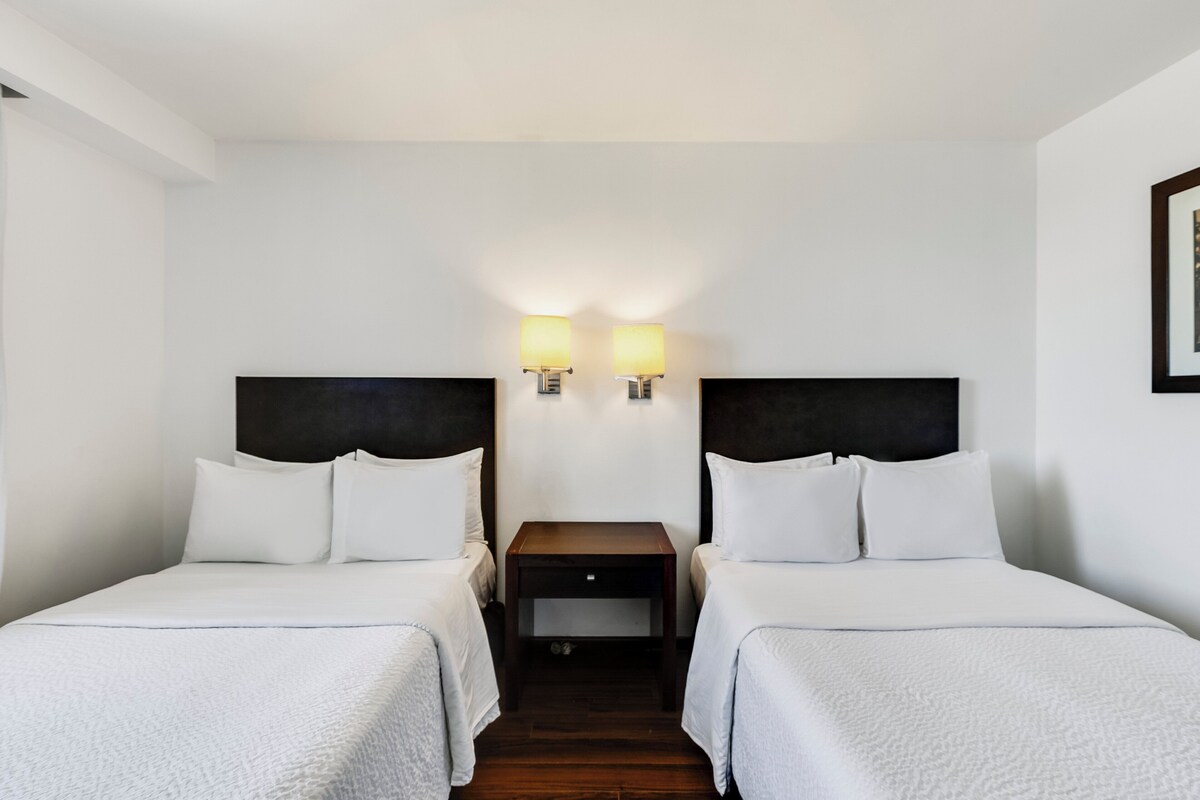 Una camera d'albergo con due letti singoli, un comodino in legno scuro posto tra essi e due lampade a parete.