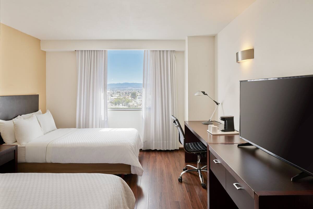 A hotel room with two beds, a desk with a TV, a chair, and a view of the city through large windows.