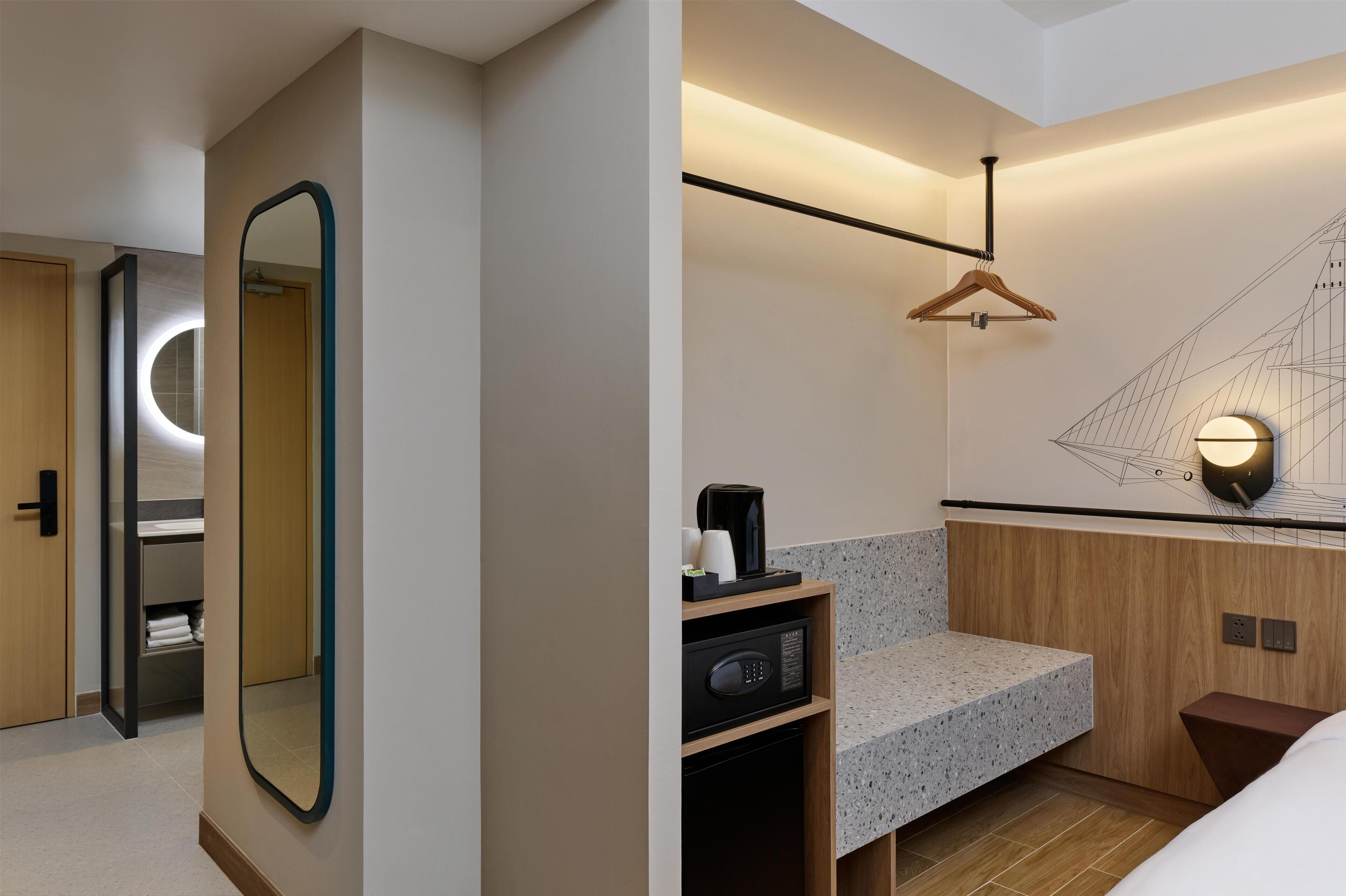 Una habitación de hotel moderna con un armario de madera, un espejo grande, una cafetera integrada y una obra de arte geométrica en la pared.