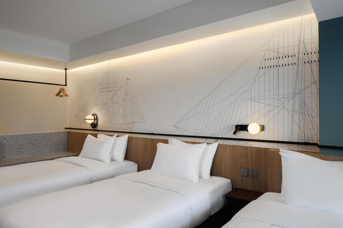 Habitación de hotel con dos camas, cabecero de madera, lámpara fijada a la pared y diseño geométrico en la pared.