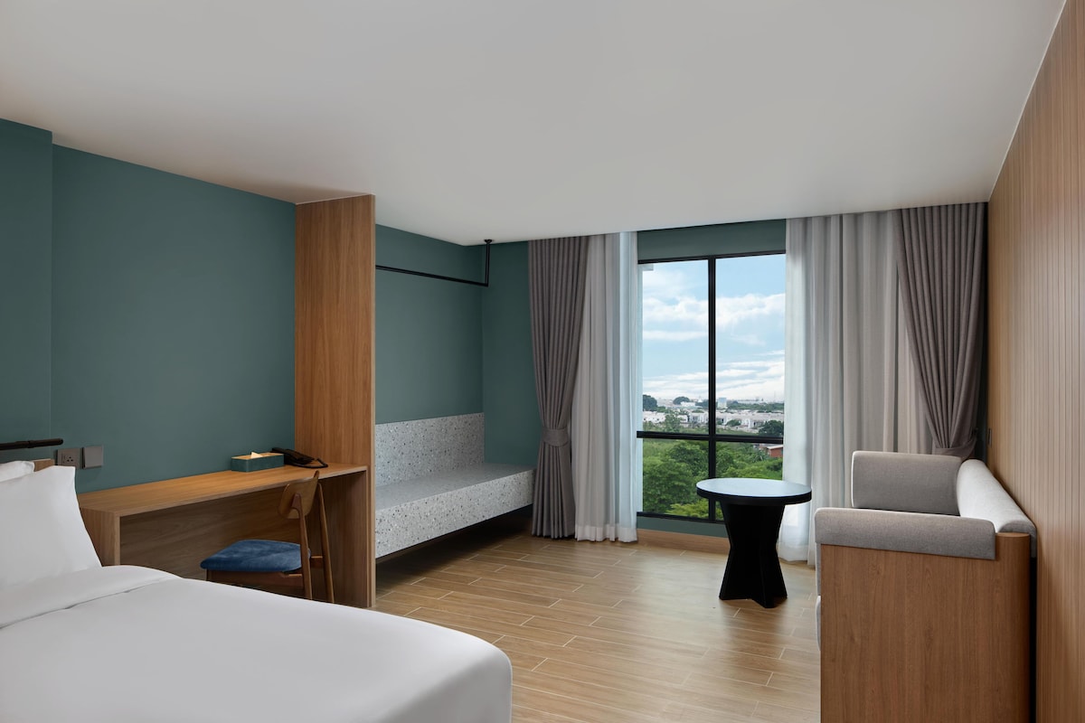 Habitación de hotel moderna con cama, escritorio, silla, mesita pequeña y balcón con cortinas.