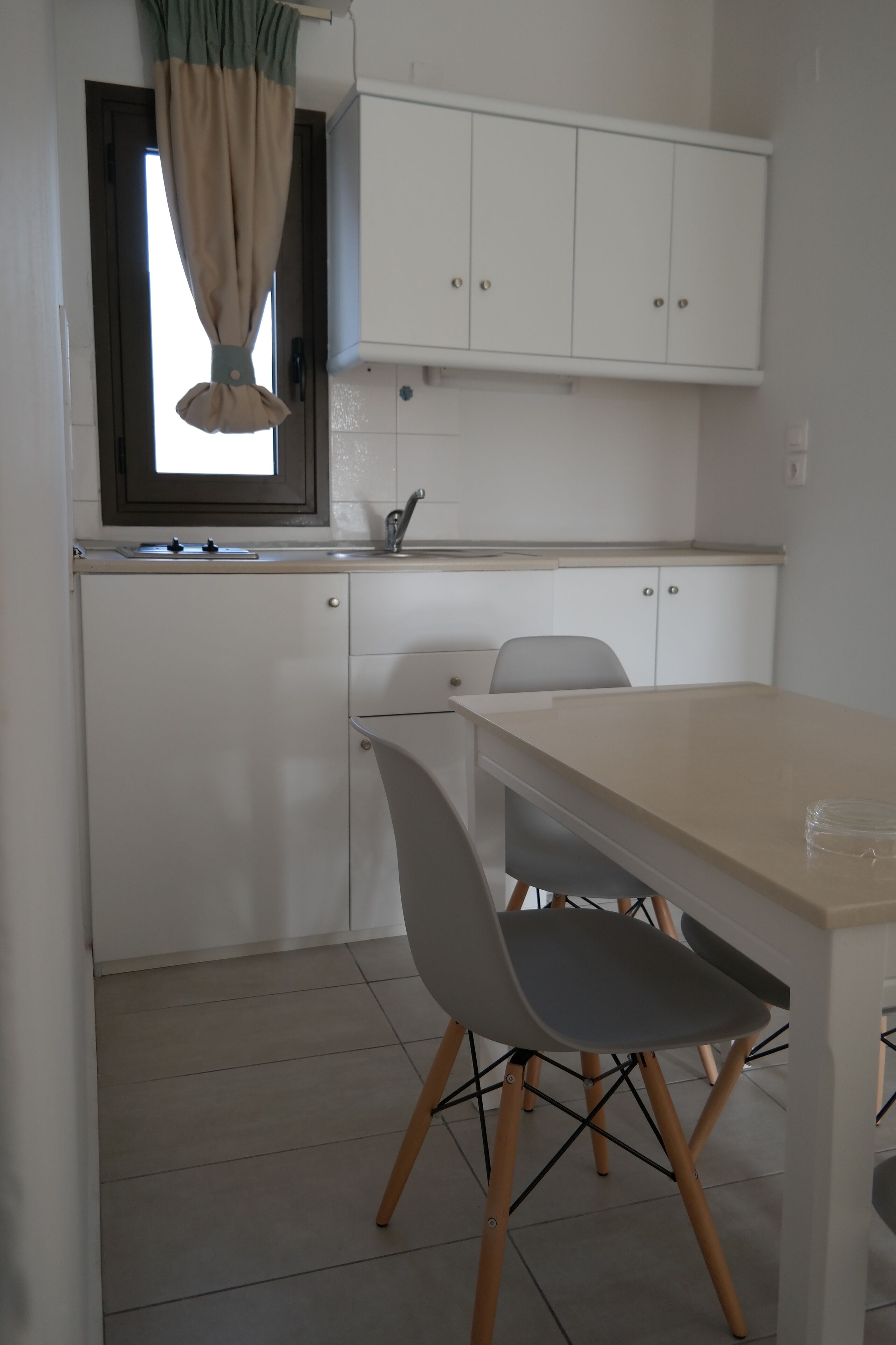 A modern kitchen with white cabinets, a window with beige curtains, and a dining area with a table and chairs.
