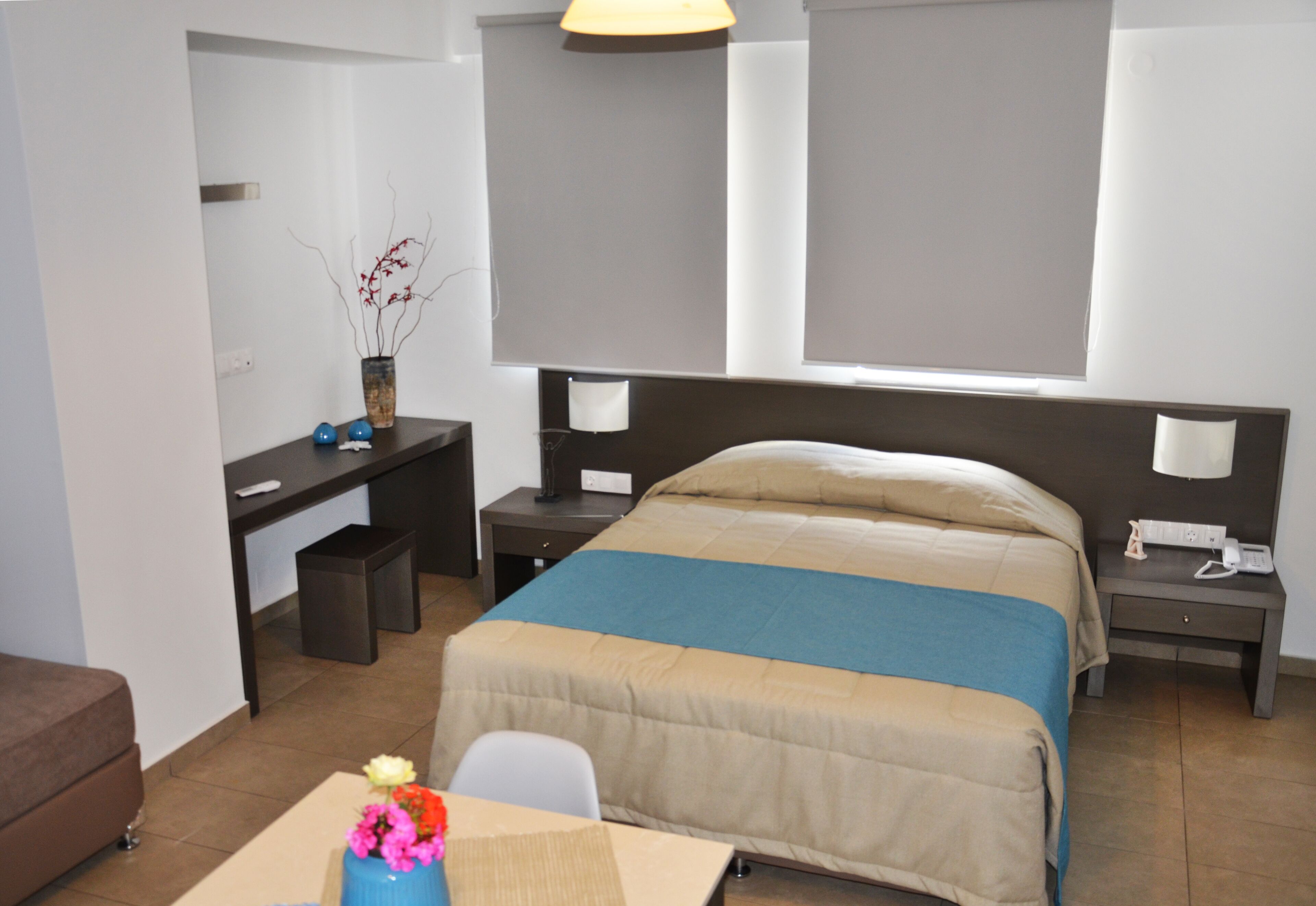 A hotel room with a bed, desk, chair, and a small table with a vase of flowers.