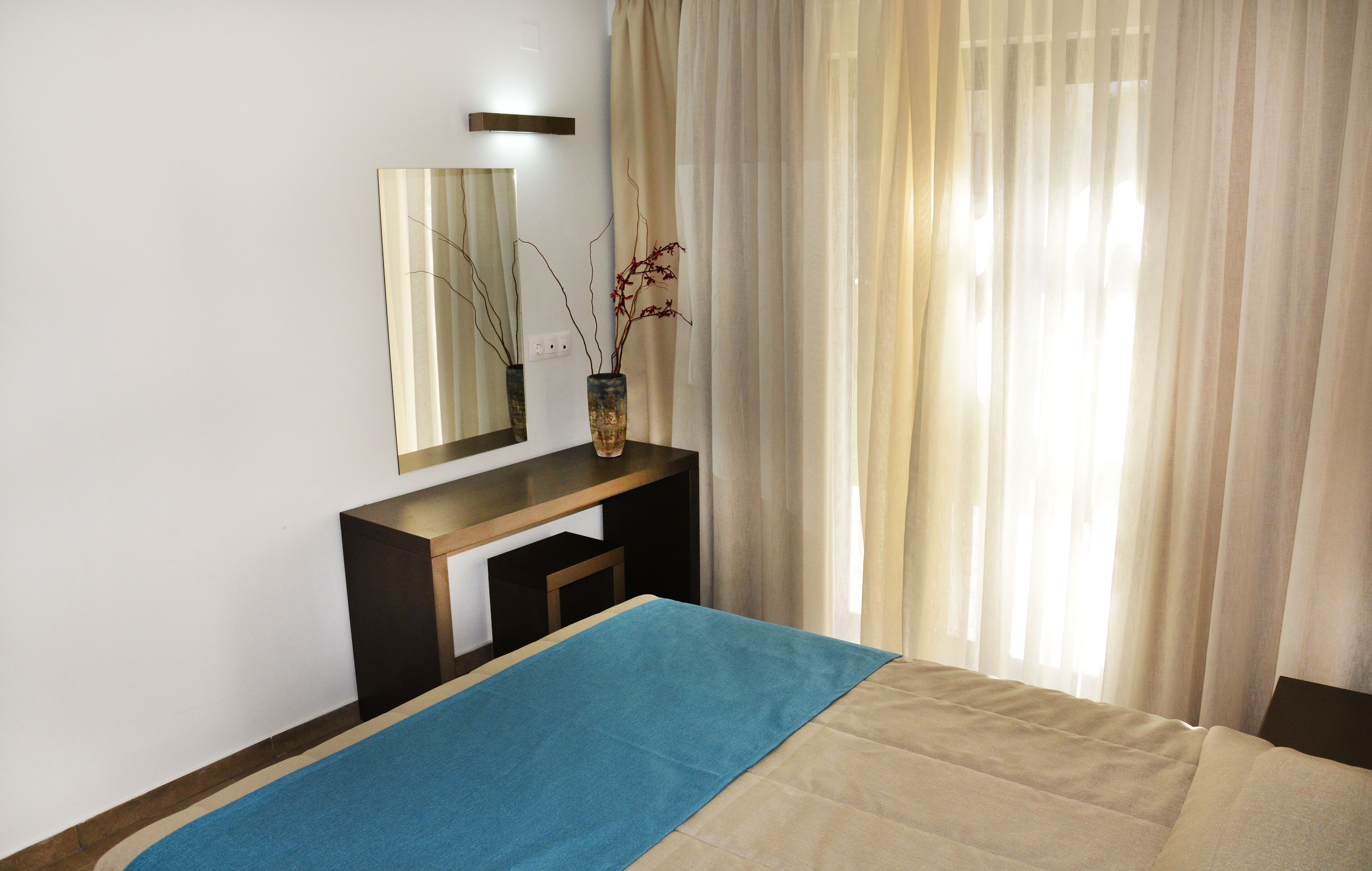 A bedroom with a bed, a bedside table with a vase, a mirror, and sheer curtains.