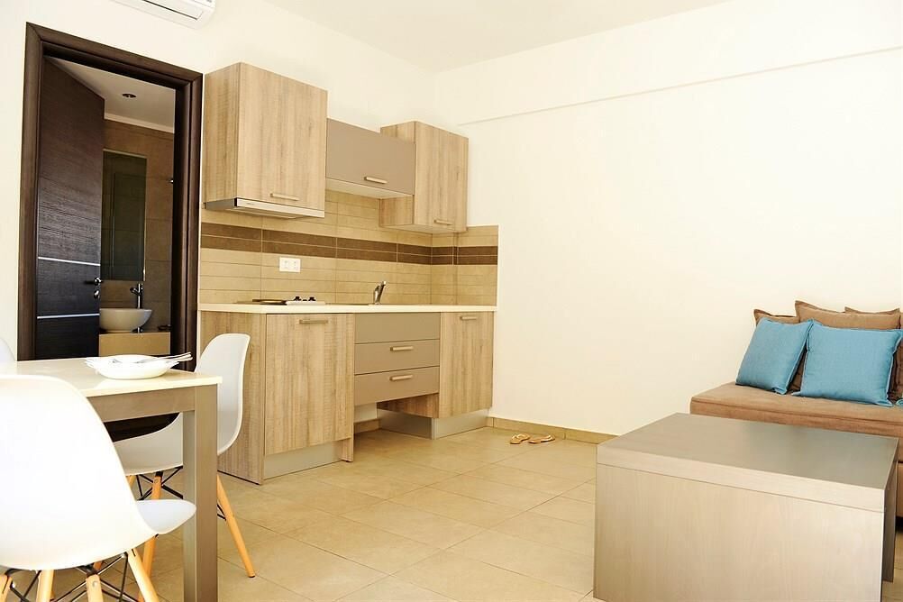 A compact kitchen with wooden cabinets, a dining area with white chairs, and a sofa with blue cushions.