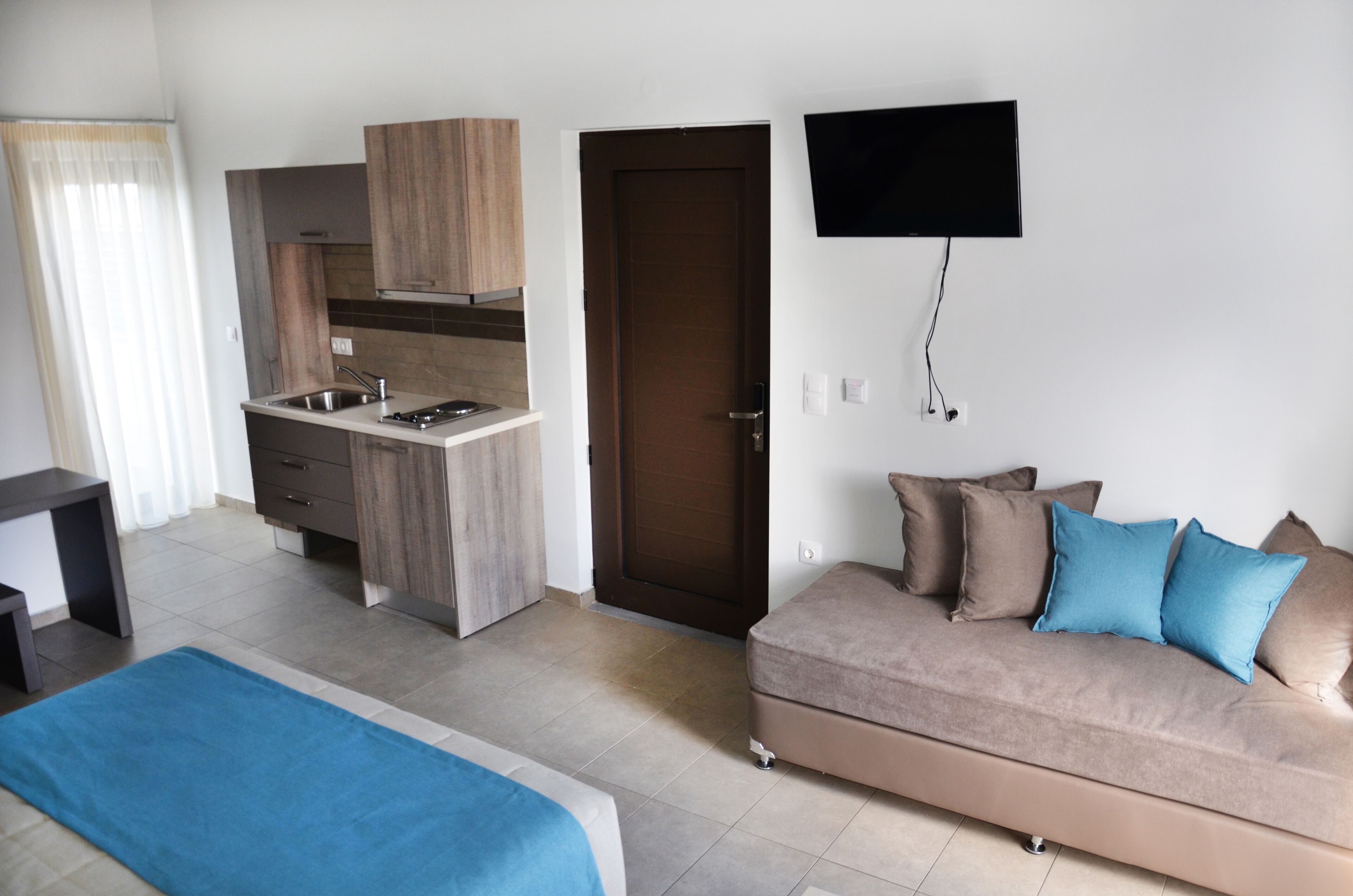 Una habitación de hotel moderna con kitchenette, un sofá con cojines azules y un televisor de pantalla plana montado en la pared.