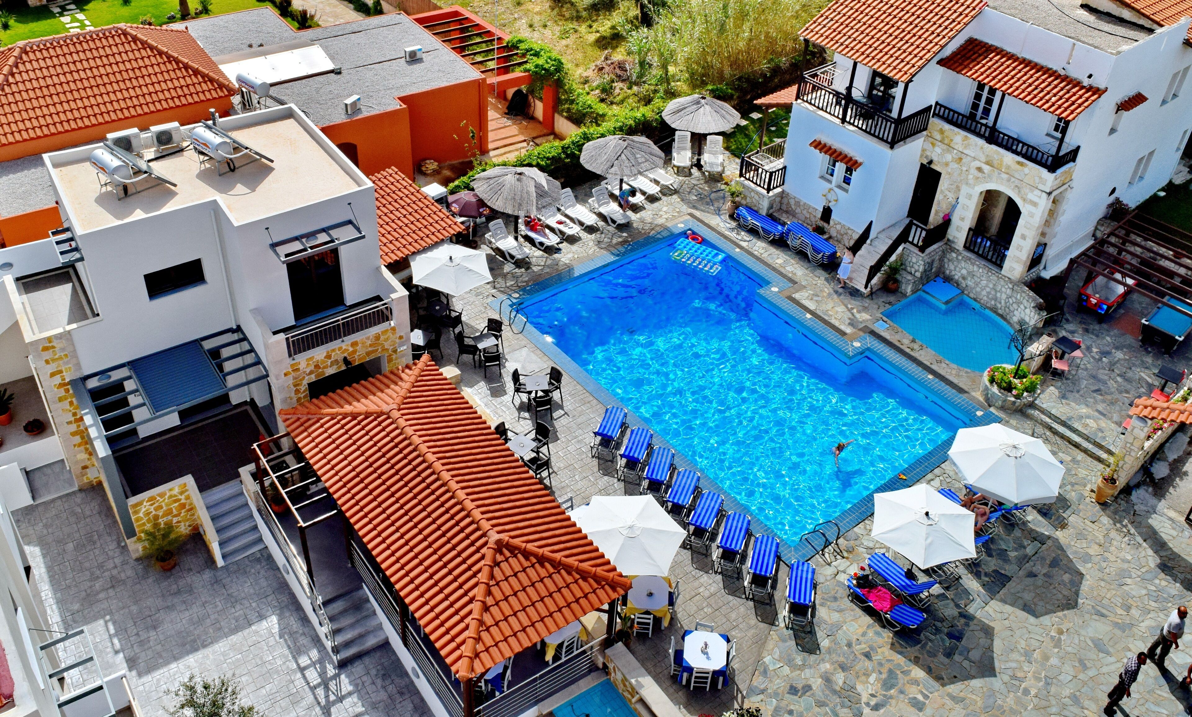 An aerial view of a hotel with a central swimming pool surrounded by lounge chairs and umbrellas, with multiple buildings and a garden area visible.