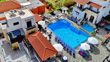 Vue aérienne d’un hôtel doté d’une piscine centrale entourée de chaises longues et de parasols, avec plusieurs bâtiments et un jardin visibles.