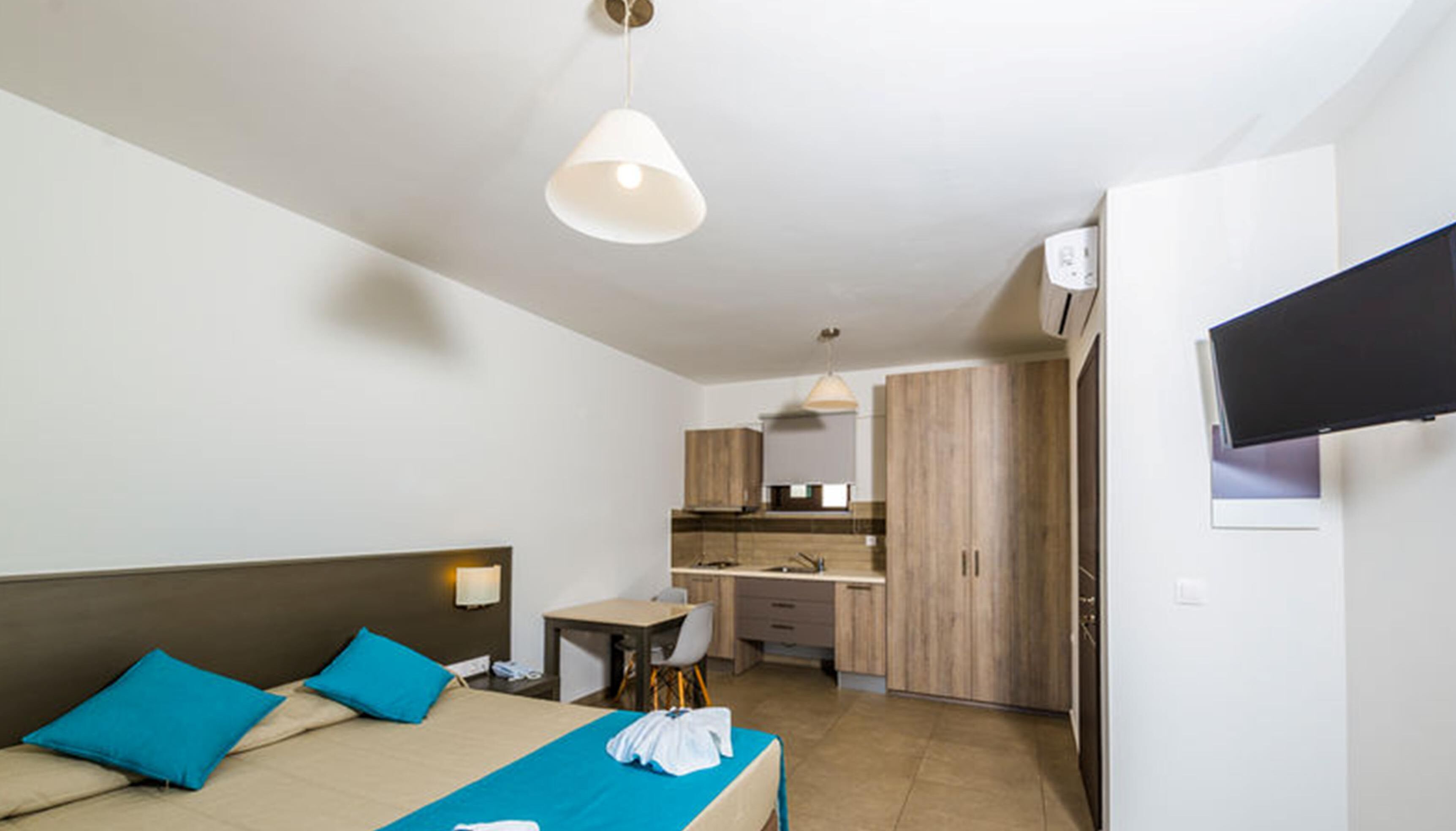 A hotel room with a bed, a desk, a kitchenette, and a TV mounted on the wall.
