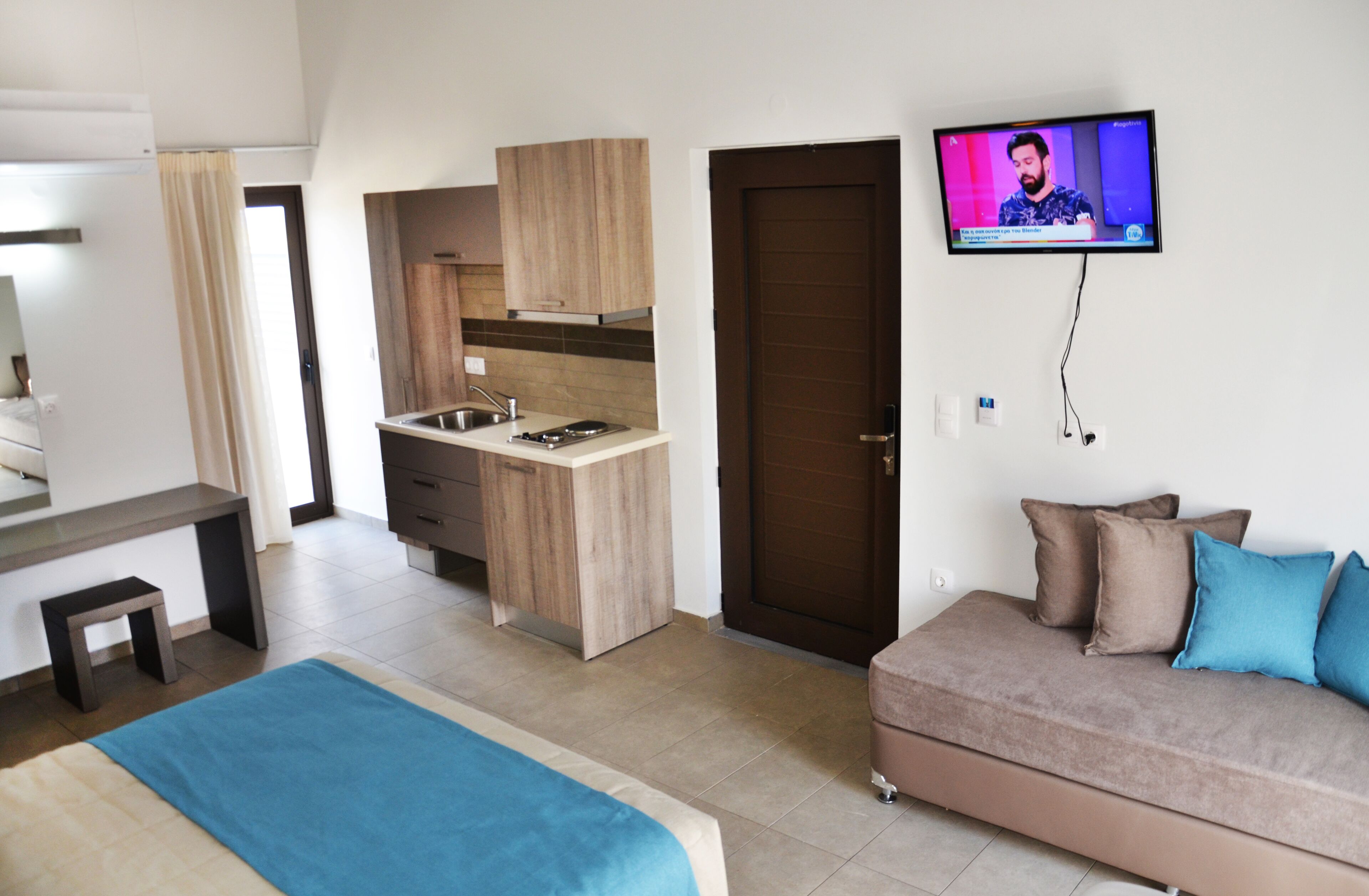 A modern hotel room with a kitchenette, a TV mounted on the wall, a sofa with cushions, and a bed with a blue blanket.