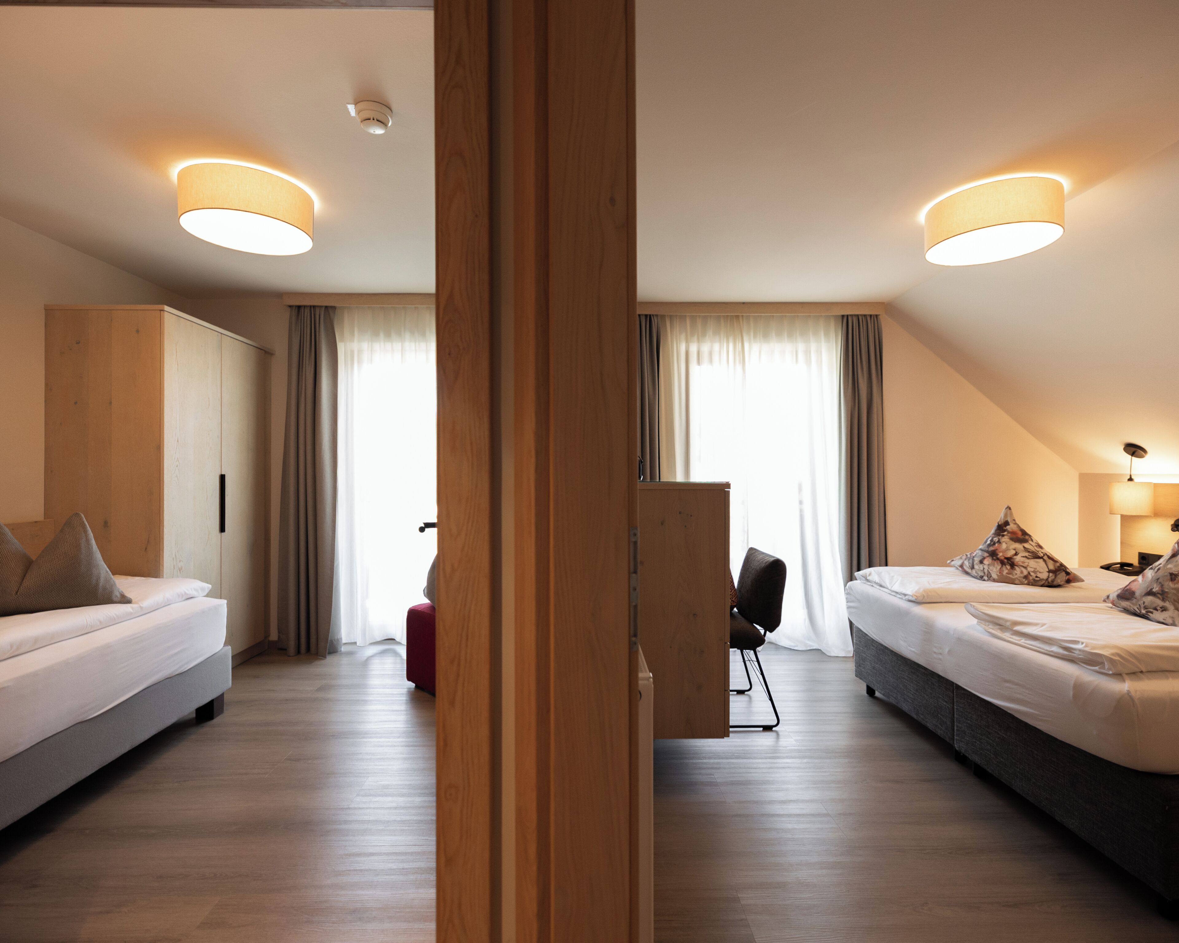 A modern hotel room with two beds, a wooden wardrobe, a desk, and a large window with curtains.