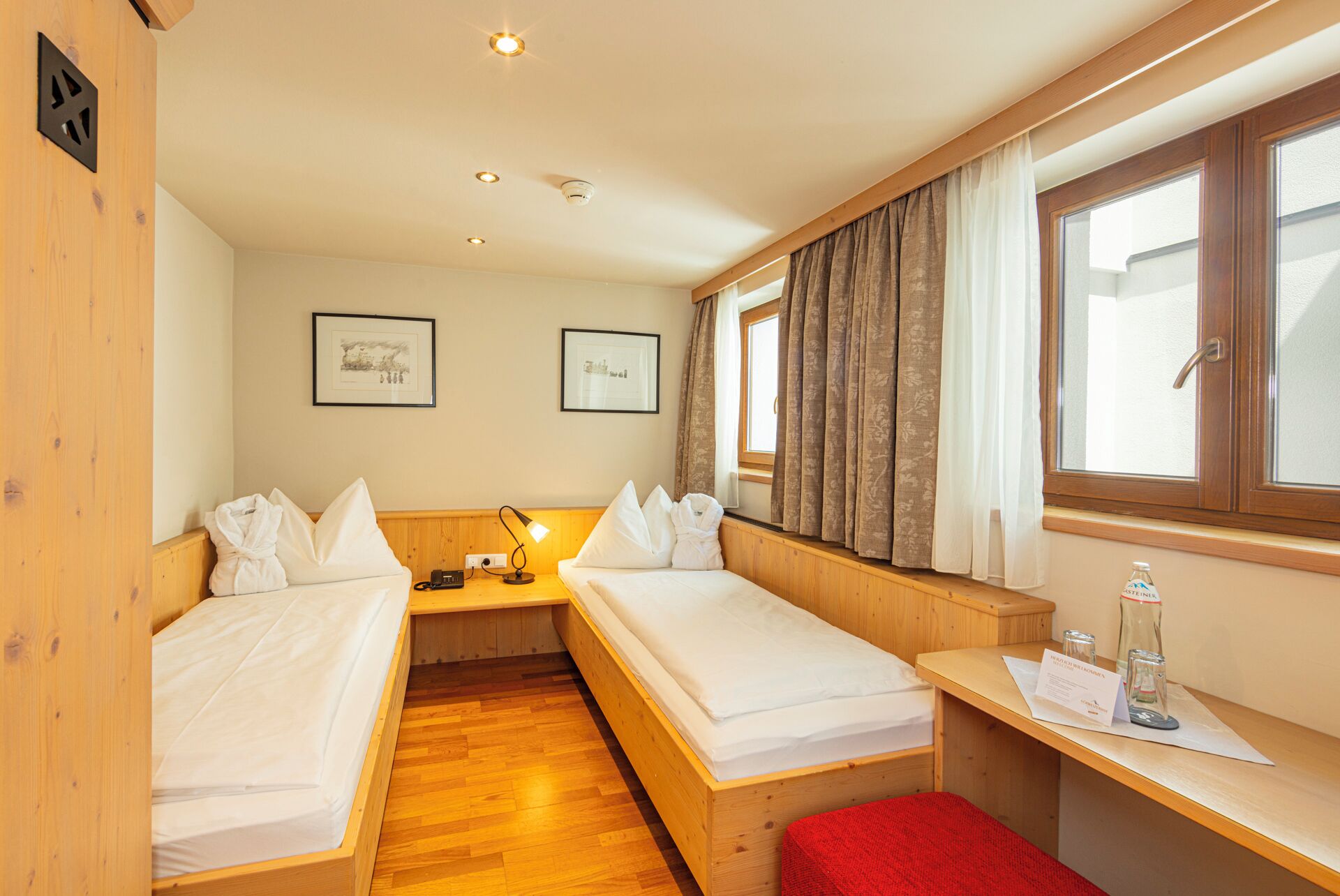 A compact hotel room with two single beds, a wooden bench, a desk with a chair, and a window with curtains.