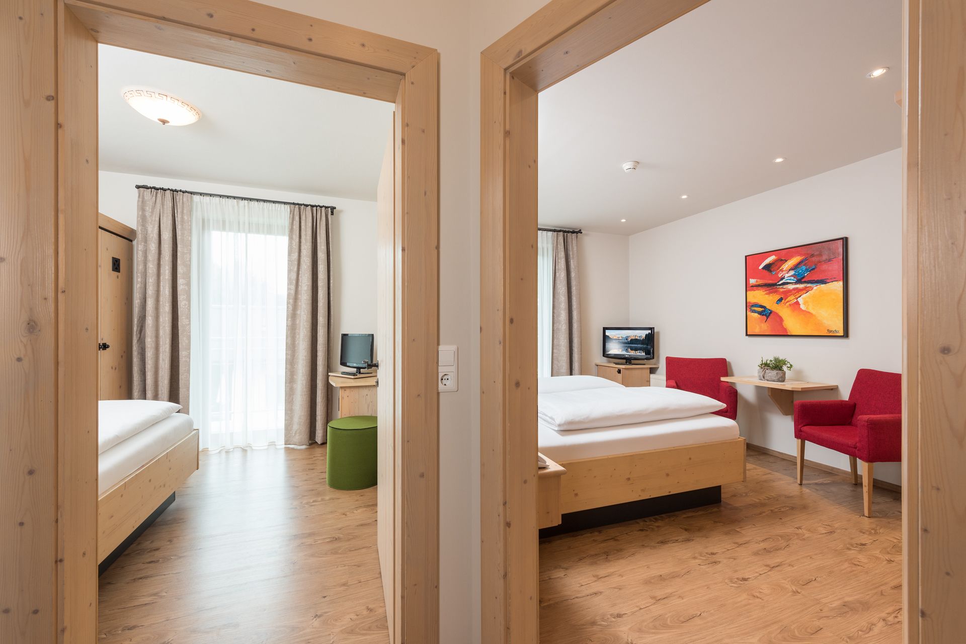 A hotel room with a wooden floor, a bed, a desk with a television, and red chairs.