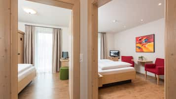 A hotel room with a wooden floor, a bed, a desk with a television, and red chairs.