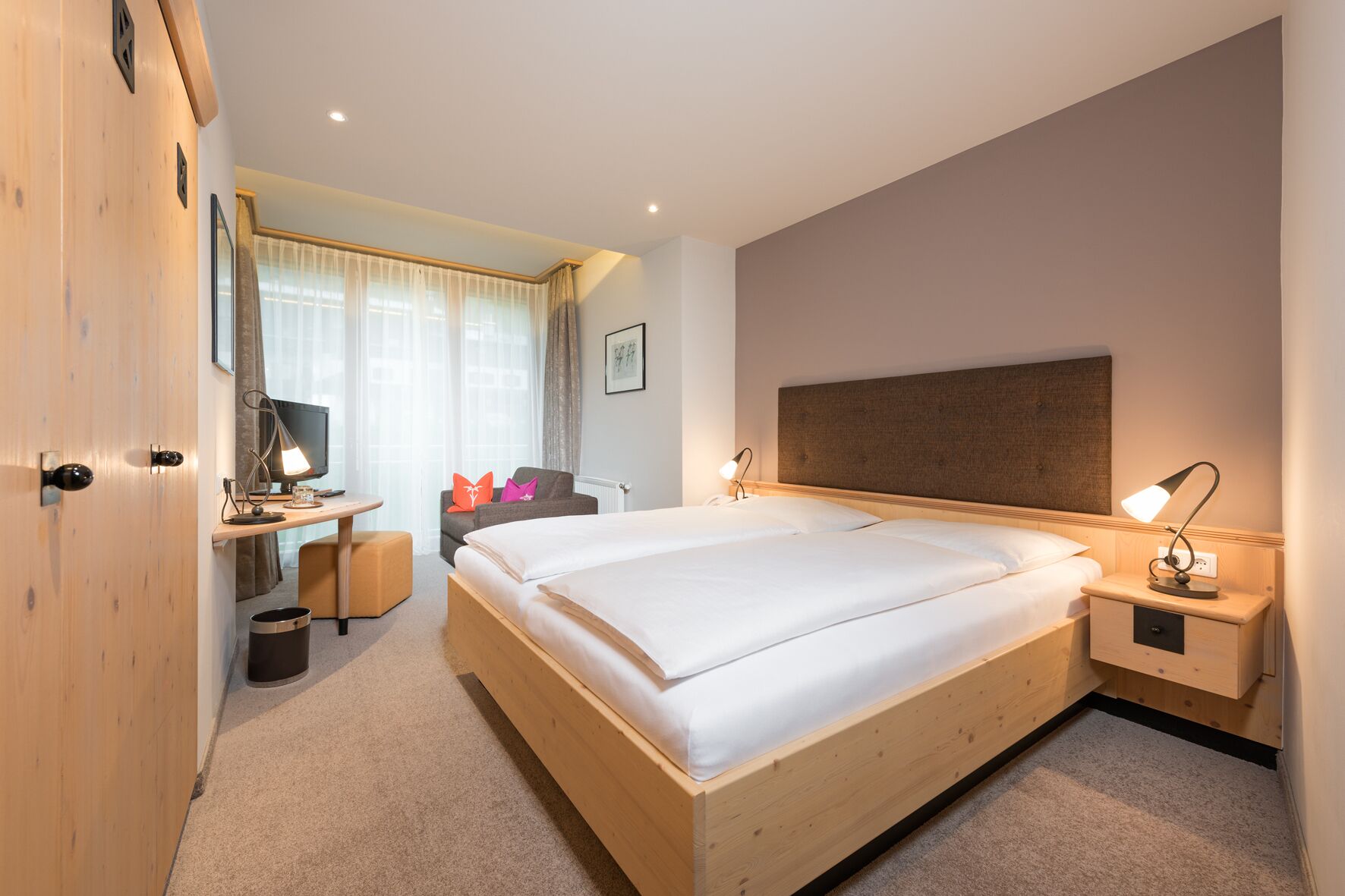 A modern hotel room with a large bed, bedside tables, a desk with a chair, a TV, and a window with curtains.