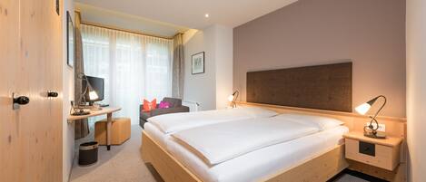 A modern hotel room with a large bed, bedside tables, a desk with a chair, a TV, and a window with curtains.