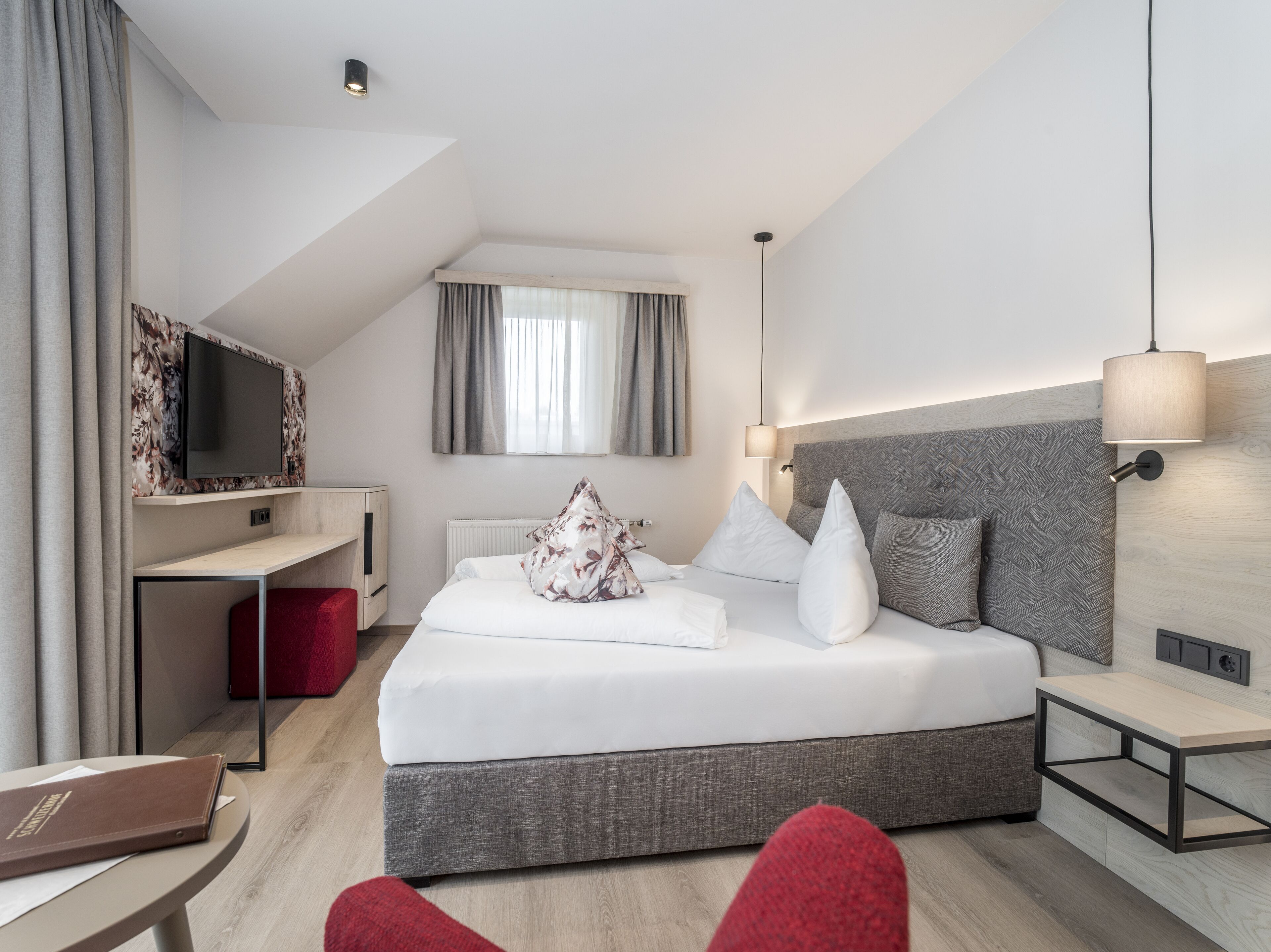 Ein modernes Hotelzimmer mit einem großen Bett, einem an der Wand montierten Fernseher, einem Schreibtisch und einem roten Sessel.