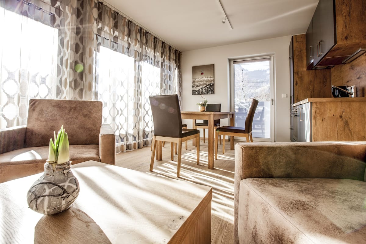 Uma sala de estar moderna com uma mesa de jantar de madeira, cadeiras e um sofá. Há um vaso com plantas verdes sobre uma mesinha lateral.