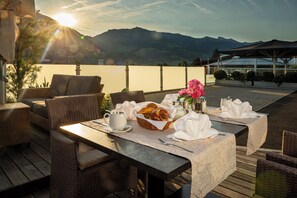 A dining table set with bread, flowers, and a pitcher on a terrace with wicker furniture and a mountain view.