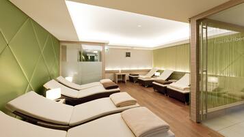 A modern spa room with multiple massage tables, a glass partition, and a visible reception area.