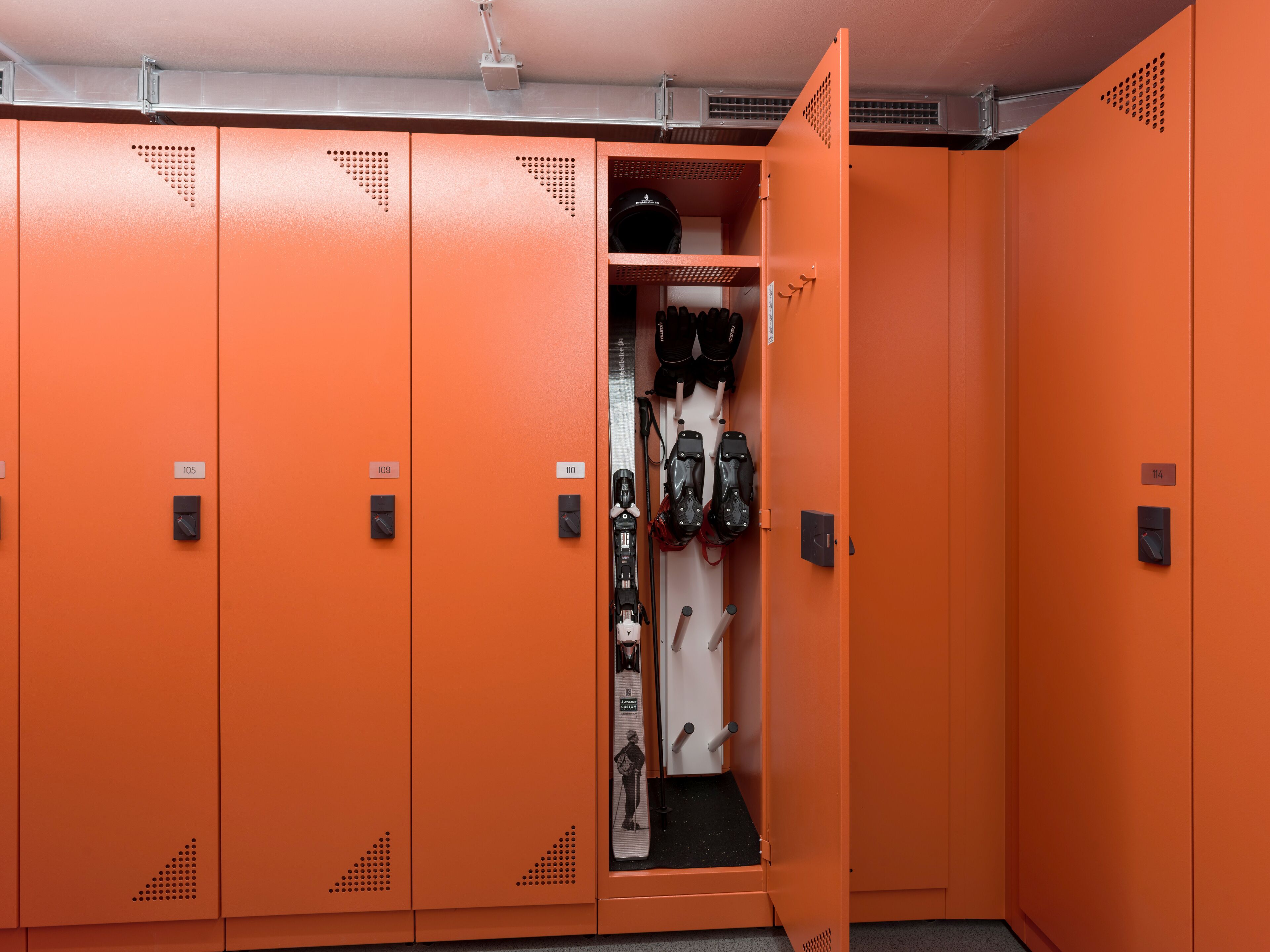 A storage room with orange lockers, some with numbers on them, and a door open to a space with shoes and a poster.
