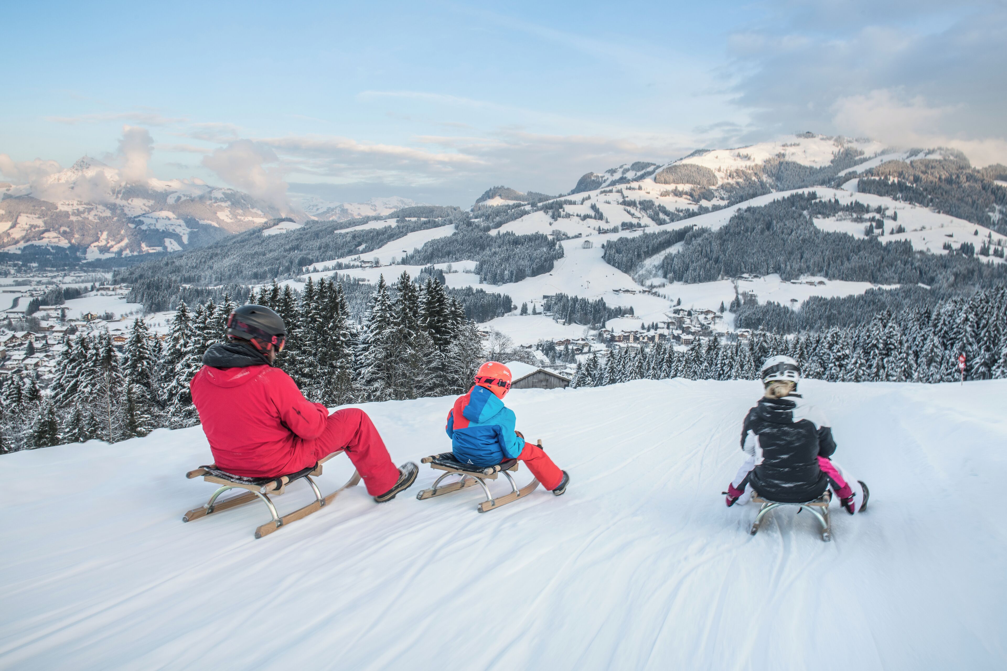 Three people sledding down a snowy slope with a mountainous landscape in the background.