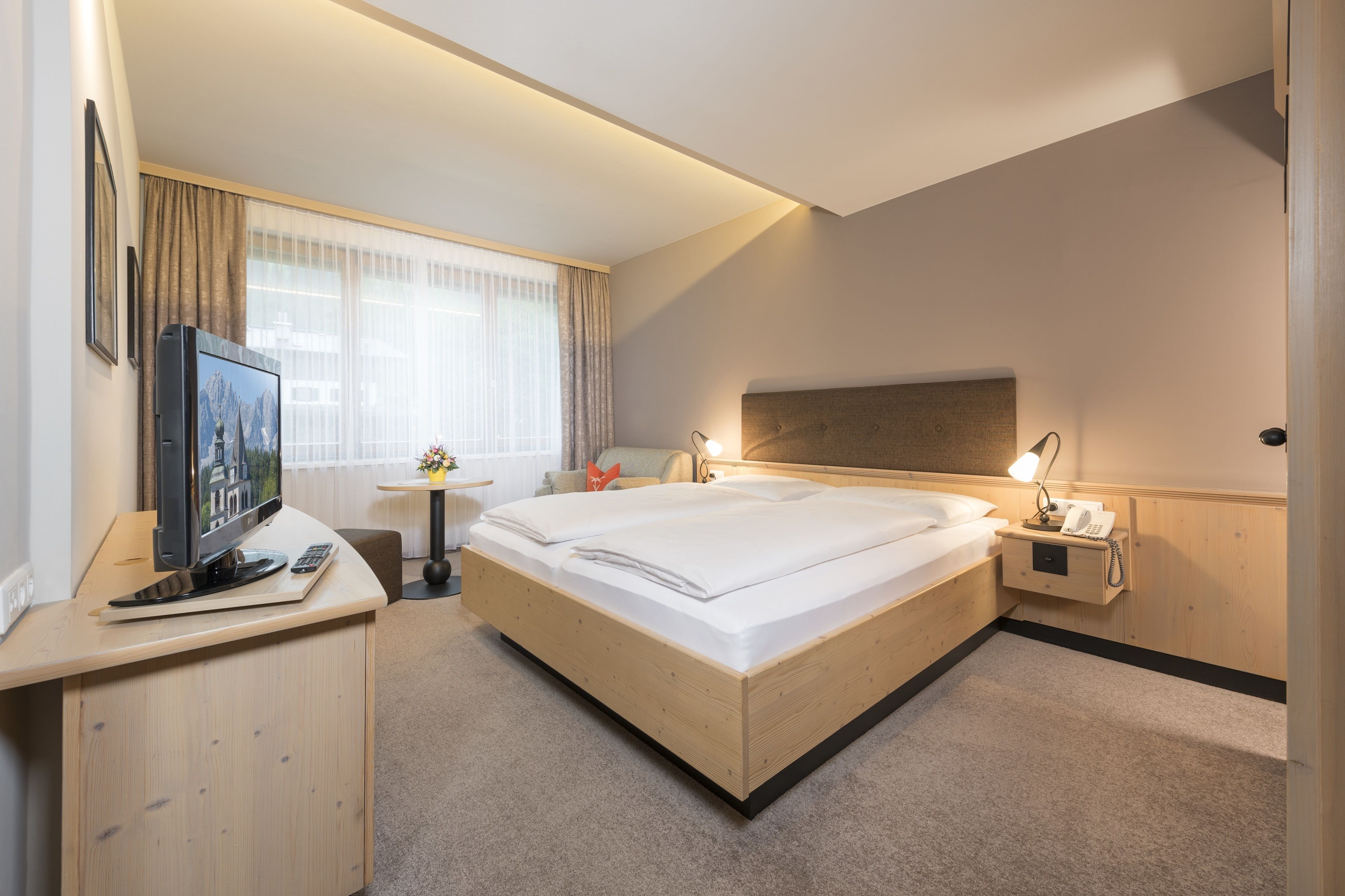 Quarto de hotel com cama de madeira, uma escrivaninha com televisão, mesa de cabeceira com abajur e janela com cortinas.