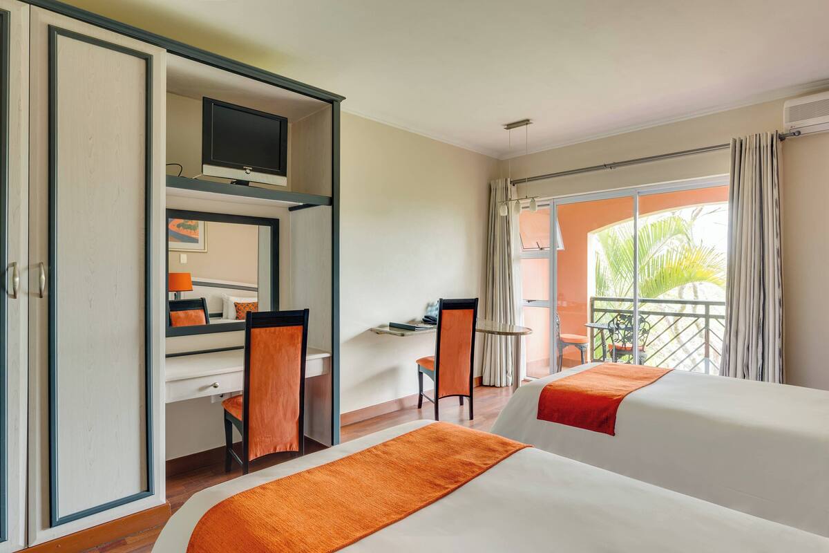 A hotel room with two beds, a desk with a chair, a television, and a balcony with a view of palm trees.