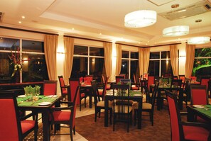 A dining area with multiple tables and chairs, large windows with curtains, and hanging light fixtures. - Protea Hotel by Marriott Umfolozi River (Mfolozi)
