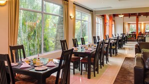 A dining area with tables set for a meal, large windows with curtains, and a view of greenery outside.
