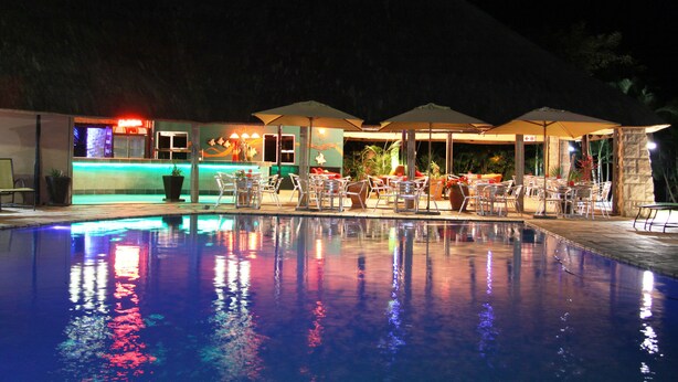 A nighttime pool area with lounge chairs and umbrellas, a bar with neon lights, and a reflection on the pool's surface.